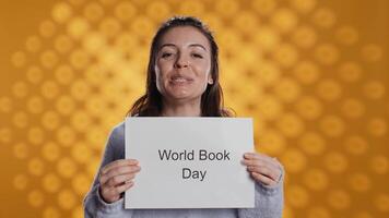 Smiling book club member holding placard with world book day message, isolated over studio background. Radiant bookworm recording promoting literacy importance, camera B video