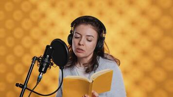 Narrator wearing headset reading aloud from book into mic against yellow background. Upbeat professional voice actor recording audiobook, creating engaging media content for listeners, camera A video