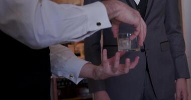 A suited man pours a drink into a glass in a relaxed setting, enjoying company with a friend. The scene exudes warmth and conviviality, capturing a moment of casual sophistication video