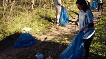 Environmental activist picking up trash with a claw tool and recycling, protecting the natural forest habitat and doing litter cleanup. Woman doing voluntary work to stop pollution. Camera B. video