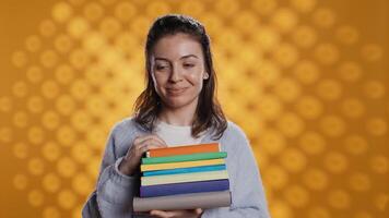 Portrait of smiling woman pointing towards empty copy spaces while holding pile of books. Radiant person with stack of novels in arms promoting reading, showing messages, studio background, camera B video