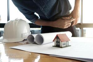 Engineer sketching a building project design with mockup house model and blueprints. Architect man working with compasses and blueprints for architectural plan. photo