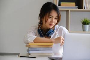 alegre joven asiático mujer utilizando ordenador portátil computadora a hogar. estudiante hembra en vivo habitación. en línea aprendiendo, estudiando , en línea compras, Lanza libre, asean concepto foto