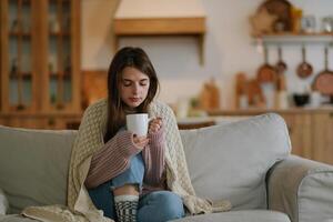 Woman freezes in winter time. Young girl wearing warm woolen socks and wrapped into plaid, holding a cup of hot drink while sitting on sofa at home. Keep warm. photo