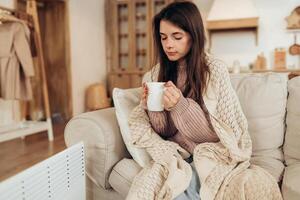 Cold home, freezing. Using heater at home in winter. Woman warming her hands sitting by device and wearing warm clothes and holding hot drink. Heating season. photo