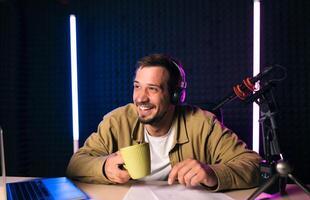 Young stylish man in mustard shirt with headphones gesturing at microphone and sharing story with audience while sitting at desk in studio with neon lighting and recording podcast photo