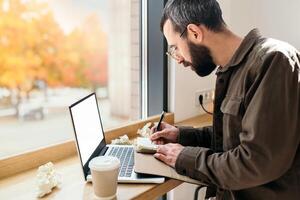 barbado negocio hombre en casual estilo escritura en un cuaderno en un cafetería. trabajo proceso, un hombre se sienta en un café con un computadora portátil, remoto trabajar, Lanza libre, otoño foto