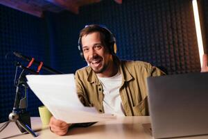 Smiling radio host with headphones reading news from paper into studio microphone at radio station with neon lights. looking at camera photo