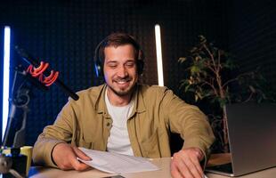 Smiling radio host with headphones reading news from paper into studio microphone at radio station with neon lights. looking at camera photo