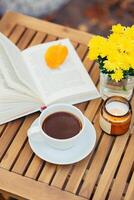 amarillo flores, taza de caliente bebida y libro en de madera mesa exterior. otoño todavía vida foto