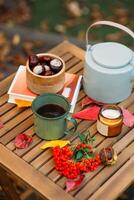 un taza de té o café y un libro en un de madera mesa en contra el antecedentes de caído hojas, otoño estación, todavía vida con hojas, un libro y viburnum y castañas en el jardín al aire libre. foto