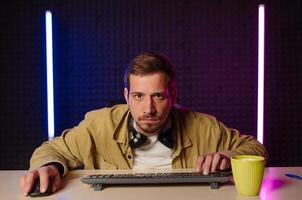 Concentrated gamer sitting at night at the computer and playing computer games with a serious face, looking at the camera. photo