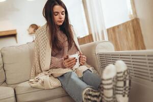 Woman freezes in winter time. Young woman with plaid near electric heater, holding a cup of hot drink while sitting on sofa at home. Keep warm. photo