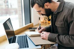 A caucasian man write in notepad on a cafe. work process. A man with a beard in a shirt is sitting in a cafe with a laptop, remote work, freelance photo