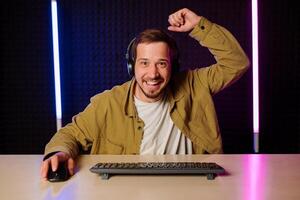 hermoso hombre en camisa ropa y auriculares sentado a un mesa en un habitación con neón luces y jugando juegos en el computadora con un sonrisa en su cara mirando a el pantalla. foto