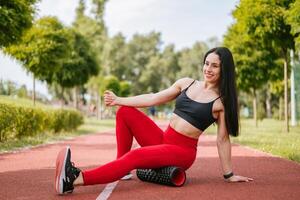 Sporty woman training on summer day, massaging and stretching hamstring and ankle leg muscles on foam roller, performing fascia exercise. Health care, workouts routine concept photo