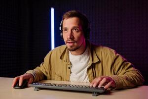 hermoso hombre en camisa ropa y auriculares sentado a un mesa en un habitación con neón luces y jugando juegos en el computadora con un sonrisa en su cara mirando a el pantalla. foto