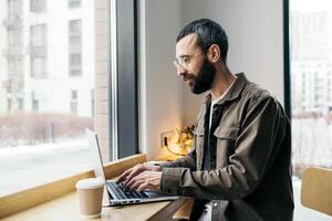 caucásico masculino persona de libre dedicación trabajando en ordenador portátil en cafetería. un hombre con un barba en un camisa se sienta en un café y bebidas café. remoto trabajo foto