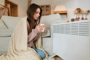 joven mujer en calentar ropa y tartán participación taza de caliente bebida mientras sentado en sofá a hogar cerca eléctrico calentador foto