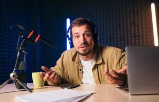 Young stylish man in mustard shirt with headphones gesturing at microphone and sharing story with audience while sitting at desk in studio with neon lighting and recording podcast photo