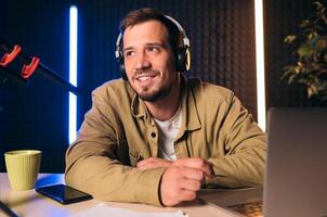 Young stylish man in mustard shirt with headphones gesturing at microphone and sharing story with audience while sitting at desk in studio with neon lighting and recording podcast photo