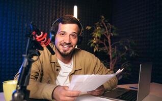 Smiling radio host with headphones reading news from paper into studio microphone at radio station with neon lights. looking at camera photo