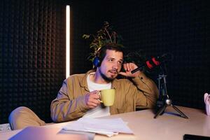 Young stylish man in mustard shirt with headphones gesturing at microphone and sharing story with audience while sitting at desk in studio with neon lighting and recording podcast photo