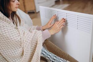 Cold home, freezing. Using heater at home in winter. Woman warming her hands sitting by device and wearing warm clothes. Heating season. photo