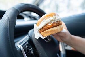 asiático dama participación hamburguesa a comer en auto, peligroso y riesgo un accidente. foto