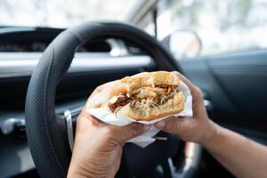 Asian lady holding hamburger and French fries to eat in car, dangerous and risk an accident. photo