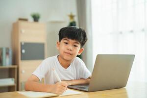 A young boy sits at a table with a laptop and a pencil. He is focused on writing something in his notebook photo