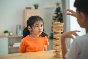 un niña en un naranja camisa es sentado a un mesa con un apilar de de madera bloques un chico es en pie detrás su, acecho su. escena es juguetón y alegre foto