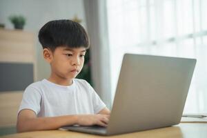 un joven chico es sentado a un mesa con un ordenador portátil en frente de a él. él es enfocado en el pantalla, posiblemente trabajando en un colegio asignación o jugando un juego foto