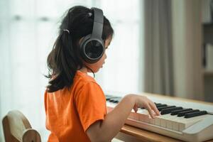 un joven niña es sentado a un piano, vistiendo auriculares. ella es vistiendo un naranja camisa. el habitación es lleno con libros y un teclado foto