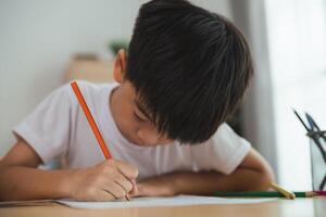 un joven chico es sentado a un escritorio y escritura con un lápiz. él es enfocado en su trabajo y él es en un grave humor. concepto de un niño aprendizaje y practicando su escritura habilidades foto