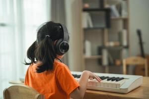 un joven niña es sentado a un piano, vistiendo auriculares. ella es vistiendo un naranja camisa. el habitación es lleno con libros y un teclado foto