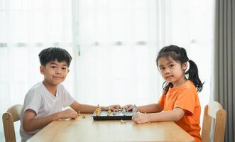 Two children are playing a game of chess. One of the children is wearing an orange shirt. The children are sitting at a wooden table photo