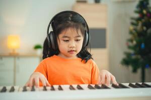 asiático joven niña es sonriente sentado a un piano, vistiendo auriculares. ella es vistiendo un naranja camisa. el habitación es lleno con libros y un teclado foto