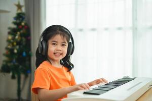 asiático joven niña es sonriente sentado a un piano, vistiendo auriculares. ella es vistiendo un naranja camisa. el habitación es lleno con libros y un teclado foto