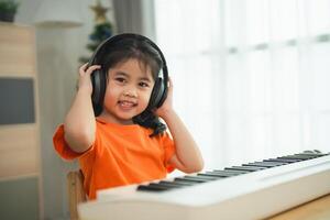 asiático joven niña es sonriente sentado a un piano, vistiendo auriculares. ella es vistiendo un naranja camisa. el habitación es lleno con libros y un teclado foto