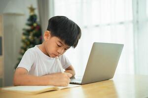 un joven chico es sentado a un mesa con un ordenador portátil y un lápiz. él es escritura alguna cosa en un pedazo de papel foto