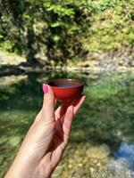 Close-up of a hand holding a traditional red tea cup against a natural water background, with focus on the cup photo