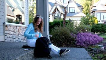 teenage girl sitting on porch in hands with phone she has white jeans blue blouse next to backpack with water private sector flowers spring summer school training waiting for parents forgot key video