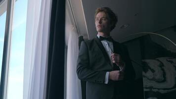 Handsome man in suit by window. Action. Gentleman in suit with bow tie looks out window. Attractive man in suit is preparing for celebration video