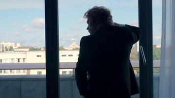 A man in a suit with long hair.Action. A handsome man stands at the window and looks at the city holding his hand behind his head, the sky is blue and small city houses are on the street. video