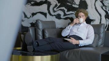 A handsome man with long hair is resting. Action.The handsome groom is sitting resting and drinking a cup of coffee in the room on the sofa. video