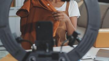 Jeune Asie Dame mode designer en utilisant mobile téléphone recevoir achat commande et montrant vêtements enregistrement vivre diffusion en ligne à magasin. petit affaires propriétaire, en ligne marché livraison concept. video