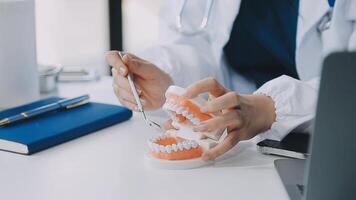 dentista sentado à mesa com modelo de dente de amostras de mandíbula e trabalhando com tablet e laptop na clínica odontológica profissional de consultório odontológico. médico trabalhando video