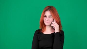 Red-haired girl talking on the phone in black clothes. Green background. bites his lips and lowers his eyes video