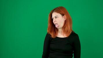 Red-haired girl in black clothes makes faces, shows tongue, smiles with different emotions green background video